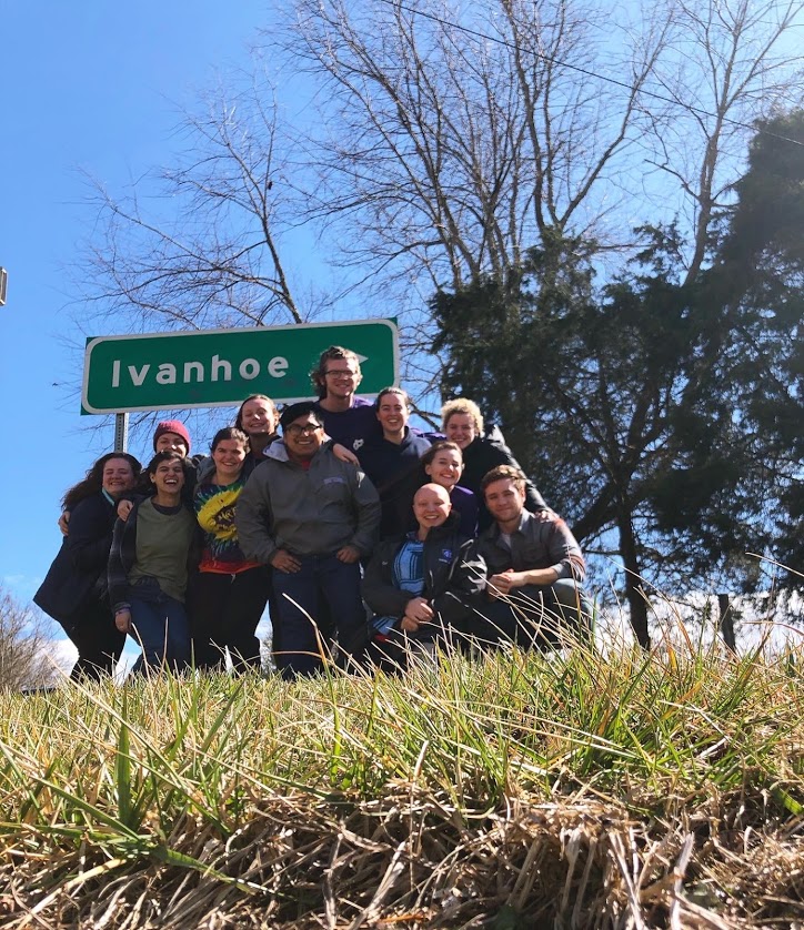 A group of twelve Holy Cross students posing in front of a sign for Ivanhoe, Virginia on a sunny day